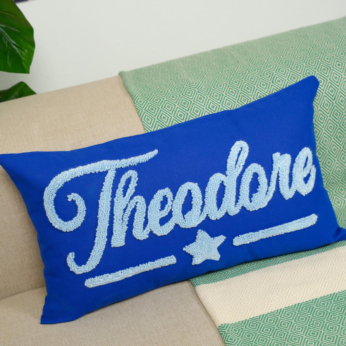 A blue throw pillow with the name "Theodore" embroidered on it, sitting on a couch with a green and white patterned blanket underneath.