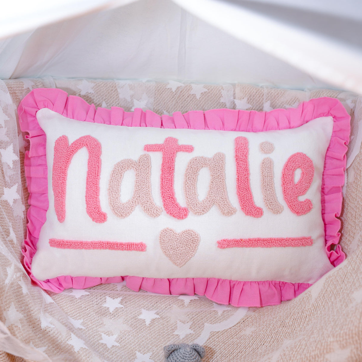 A pink and white pillow with the name "NATALIE" embroidered on it, placed on a bed with a white and beige patterned blanket.