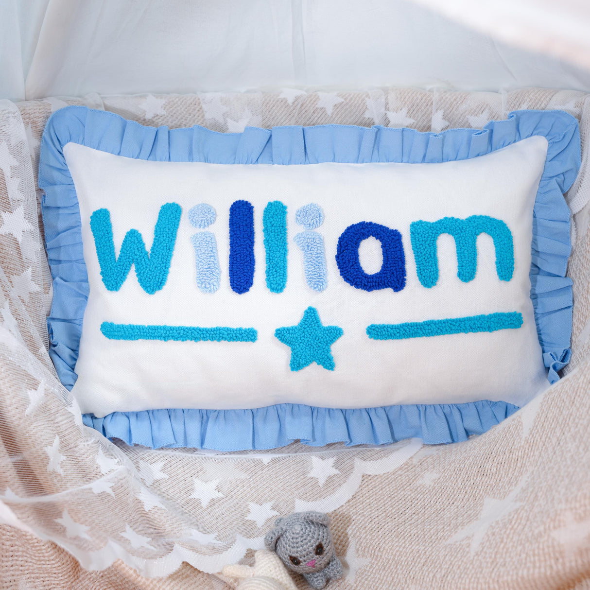 A handmade pillow with the name "William" embroidered on it, placed on a bed with a white lace blanket and a small crocheted teddy bear.