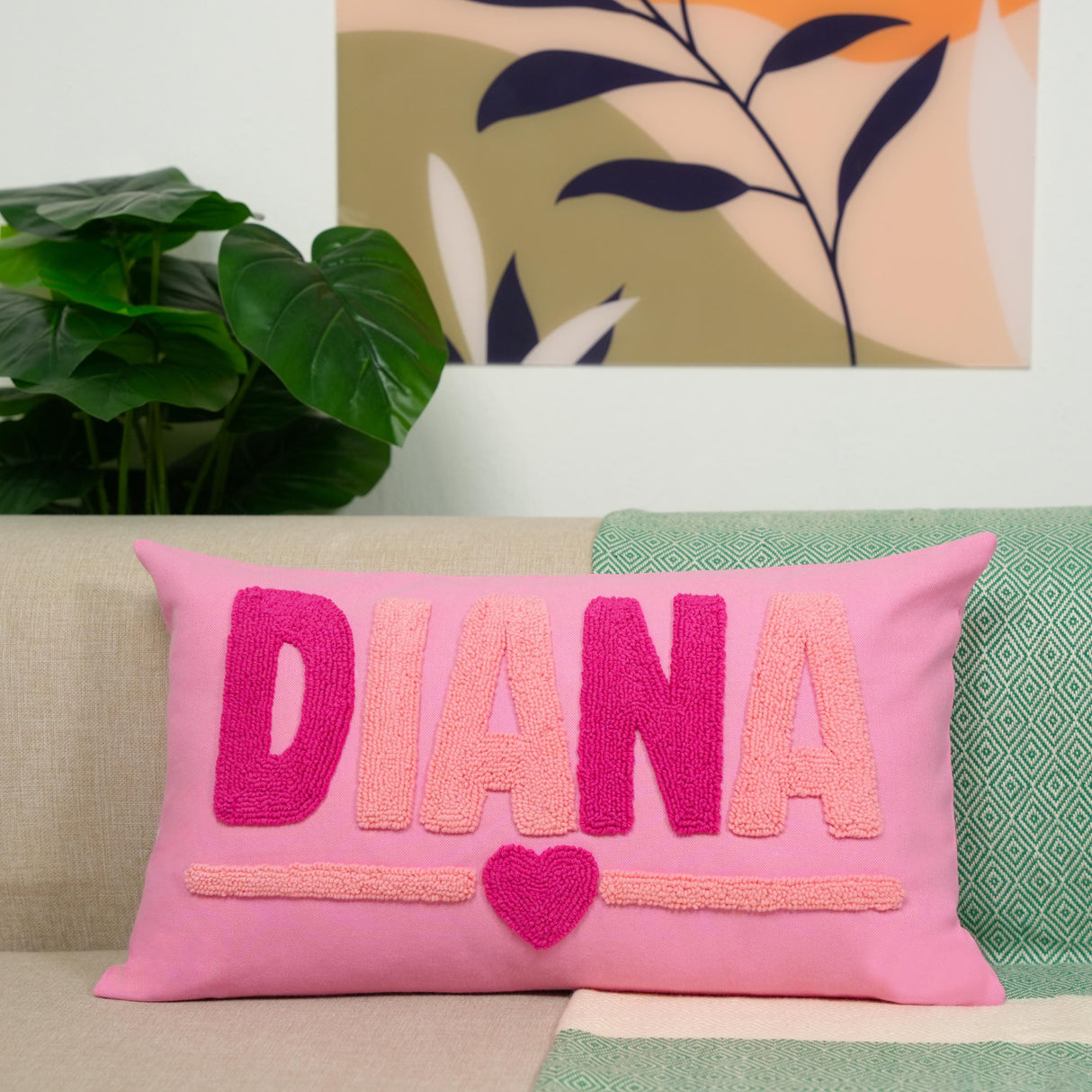 A pink throw pillow with the name "DIANA" and a heart symbol is placed on a couch, with a plant and a colorful abstract painting in the background.