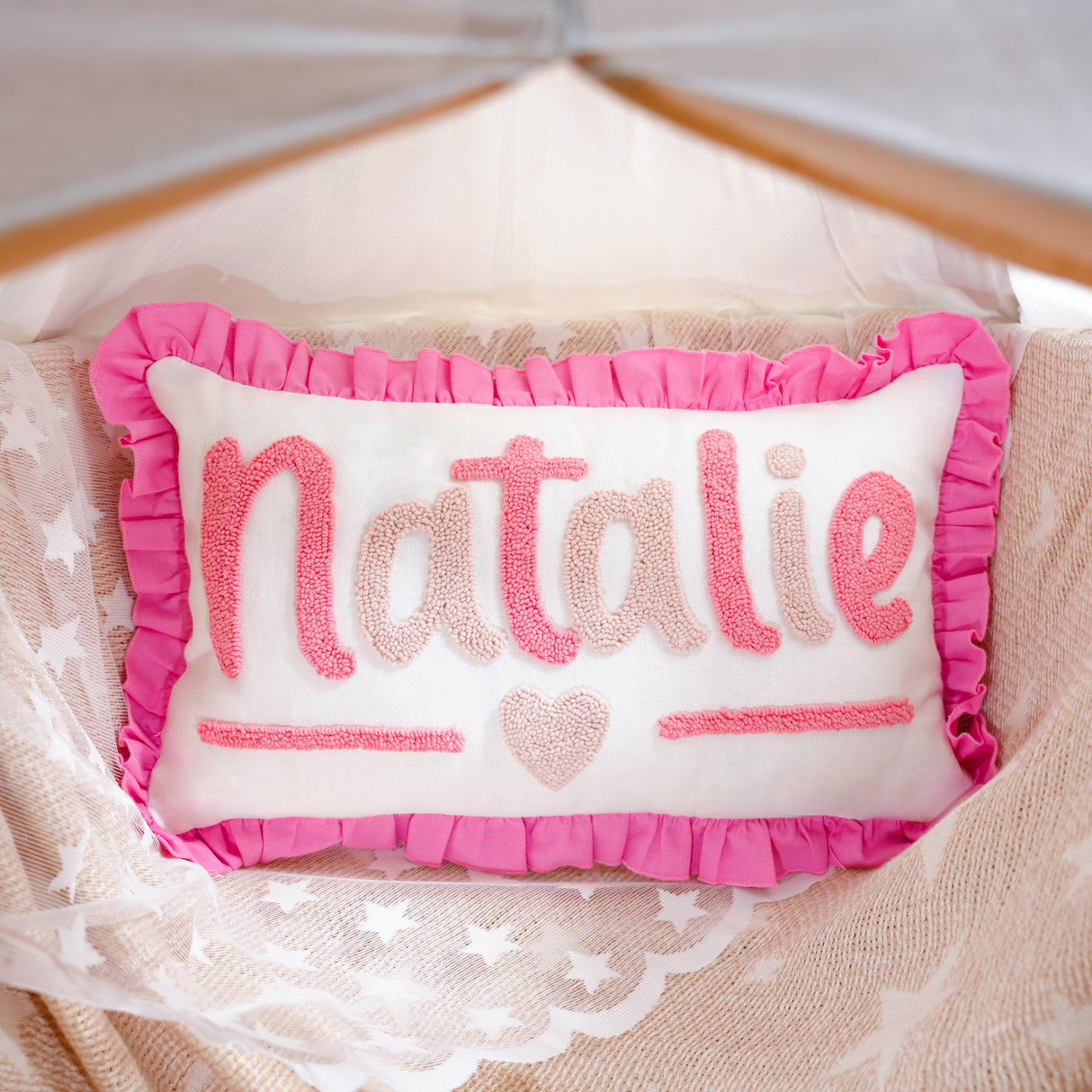 A pink and white decorative pillow with the name "NATALIE" written on it, placed on a bed with a white lace blanket.