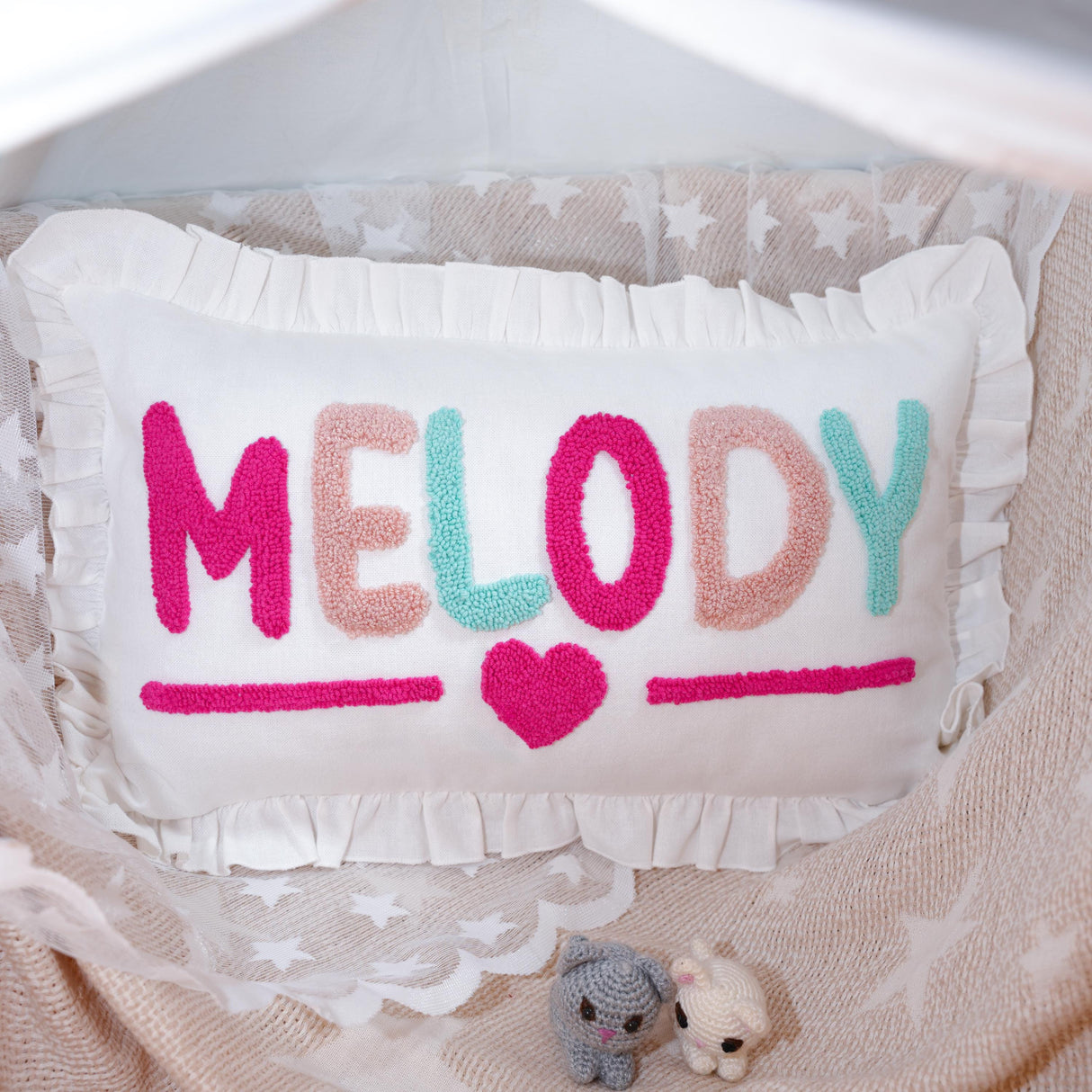 A handmade pillow with the word "Melody" written on it, featuring a heart symbol and two small crocheted animals.