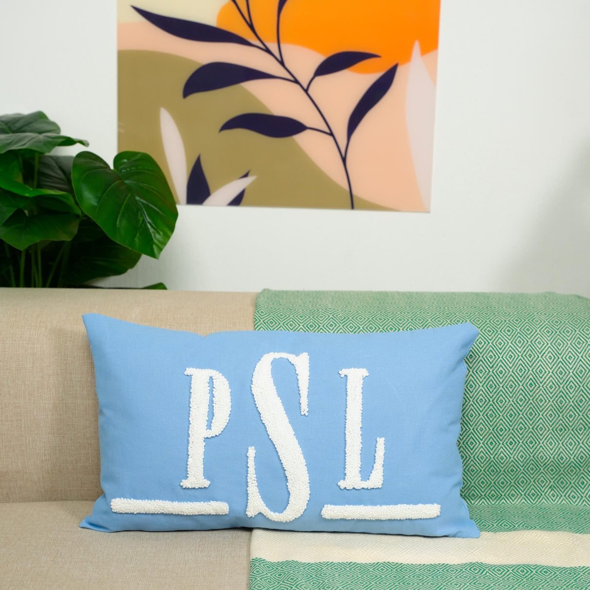 a blue pillow with the initials "PSL" on it, placed on a couch. The pillow is positioned in front of a wall with a colorful abstract painting hanging above it.