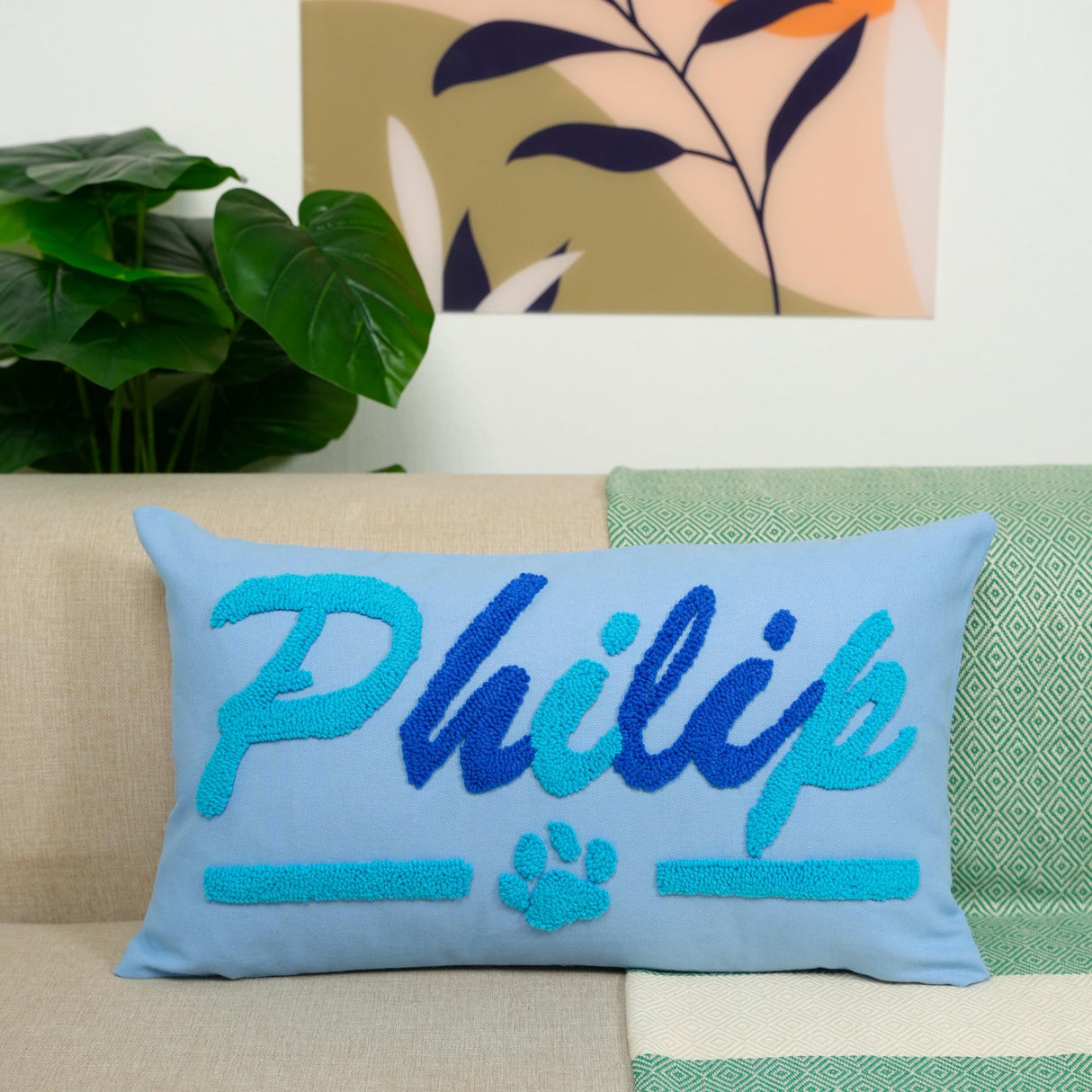 A blue throw pillow with the name "Philip" embroidered on it, placed on a couch in a room with a plant and a colorful abstract painting on the wall.