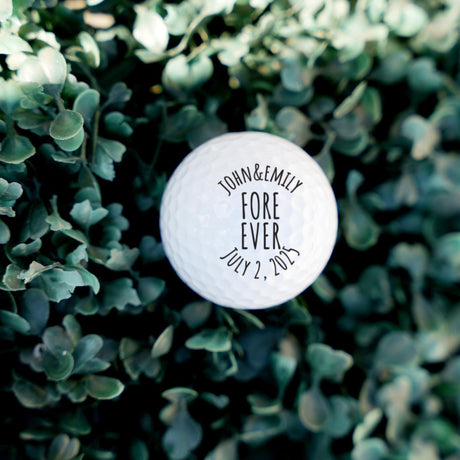 Personalized golf ball with 'John & Emily FORE EVER JULY 2, 2023' text on a green leafy background