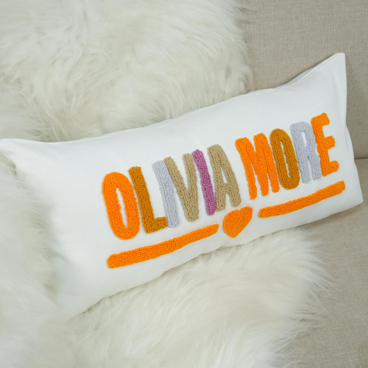 A white pillow with the word "OLIVIA MORE" embroidered on it in orange and purple colors.
