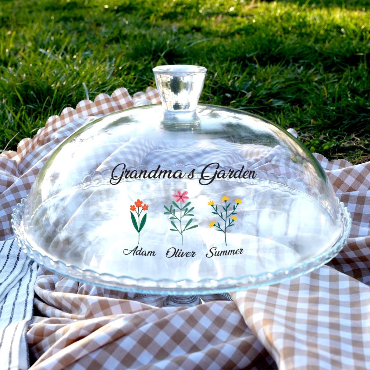 A clear glass cake stand with a decorative design, placed on a checkered cloth on a grassy lawn.