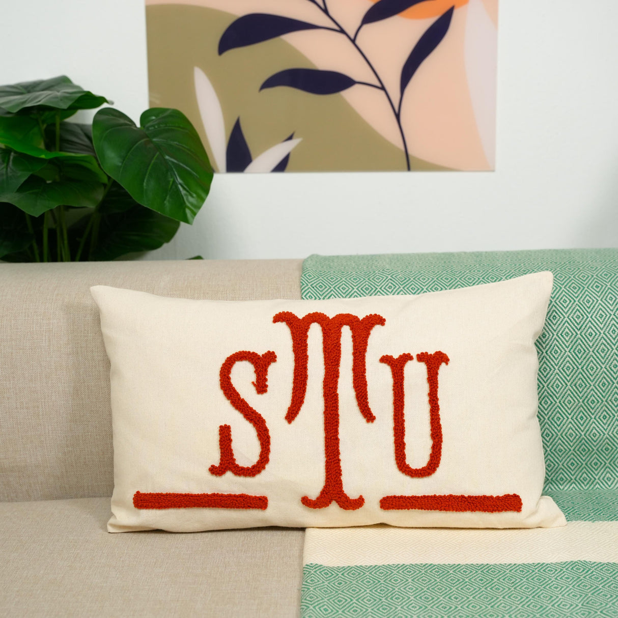 a beige couch with a white pillow adorned with red embroidered text, which reads "STU". The pillow is placed on a green and white patterned blanket. In the background, there is a plant and a framed artwork on the wall.