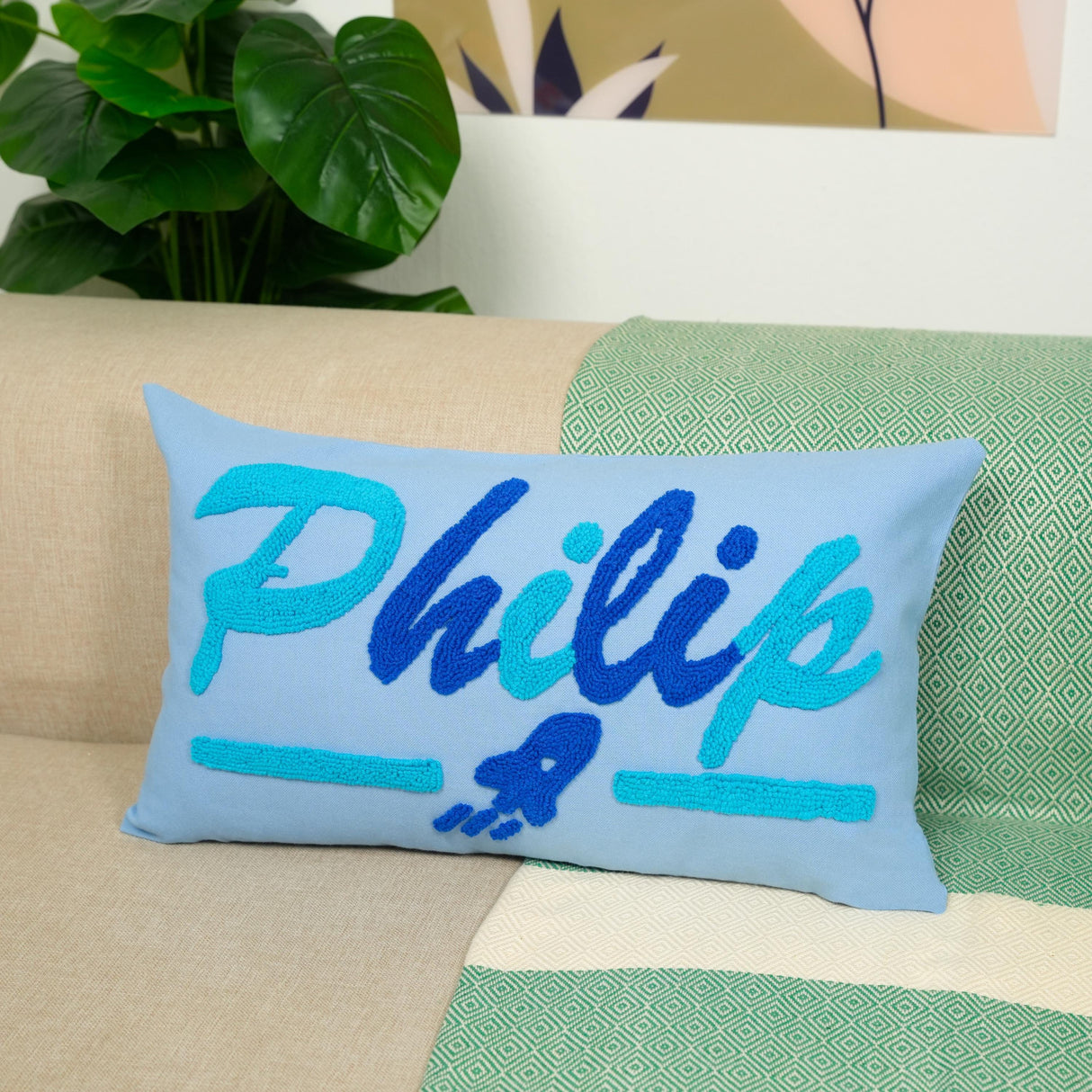 A blue and teal throw pillow with the name "Philip" embroidered on it, placed on a couch with a green and white patterned blanket underneath.
