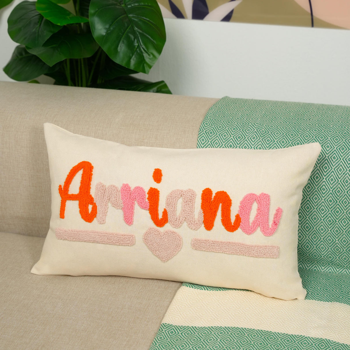 A white pillow with the name "Arriana" embroidered on it, sitting on a couch with a green and white patterned blanket underneath.