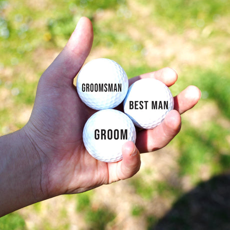 Hand holding three golf balls labeled 'Groom', 'Best Man', and 'Groomsman' with a blurred grassy background.