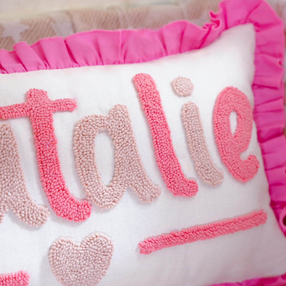 A close-up image of a decorative pillow with pink and white embroidered text and a ruffled edge.