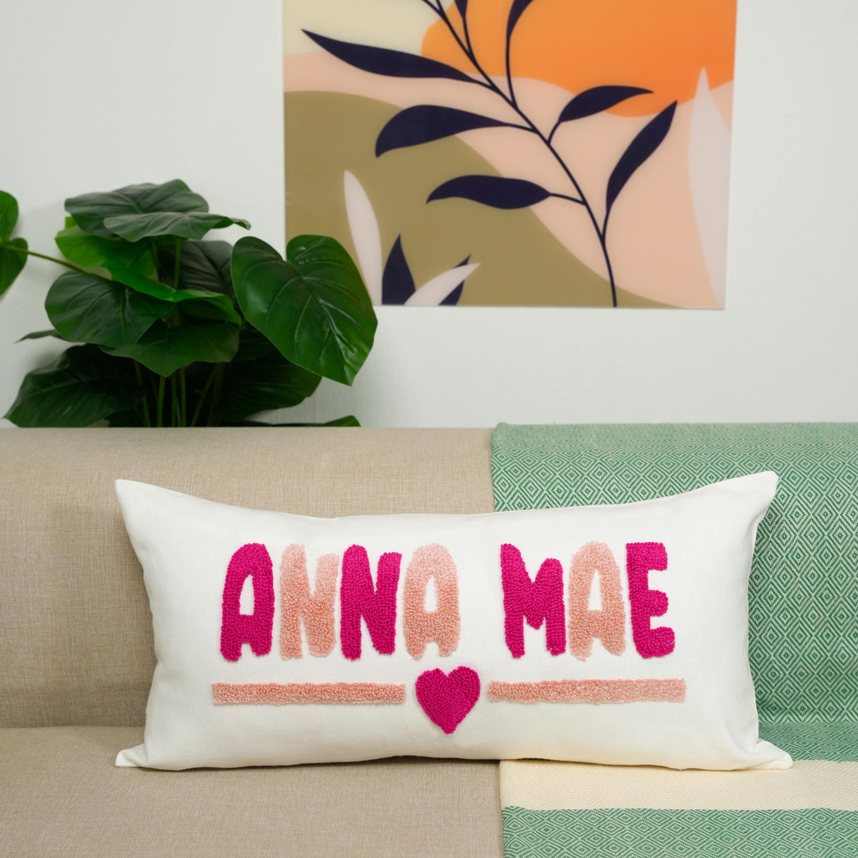 A white pillow with pink and orange text that says "ANNA MAE" and a heart symbol, placed on a couch.