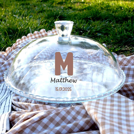 A clear glass cake stand with a glass lid, placed on a checkered cloth, with a personalized message printed on it.