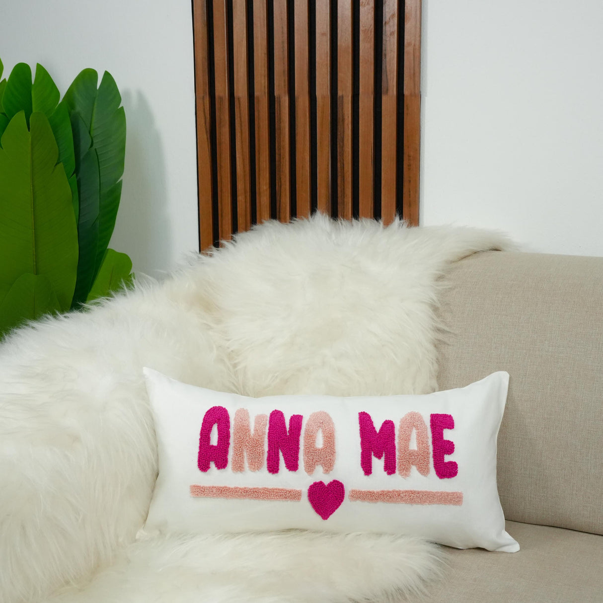 A white pillow with pink and purple embroidered text and a heart symbol sits on a beige couch, with a large green leaf plant in the background.