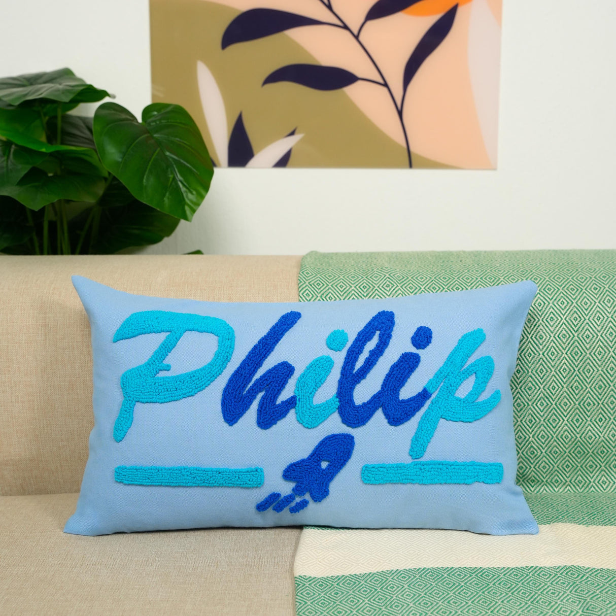 A blue and teal throw pillow with the name "Philip" embroidered on it, placed on a couch with a green and white patterned blanket underneath.