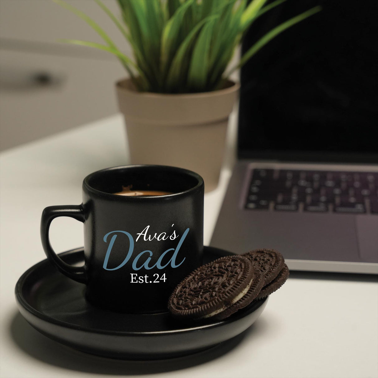 a cup of coffee with a cookie on a plate next to a laptop