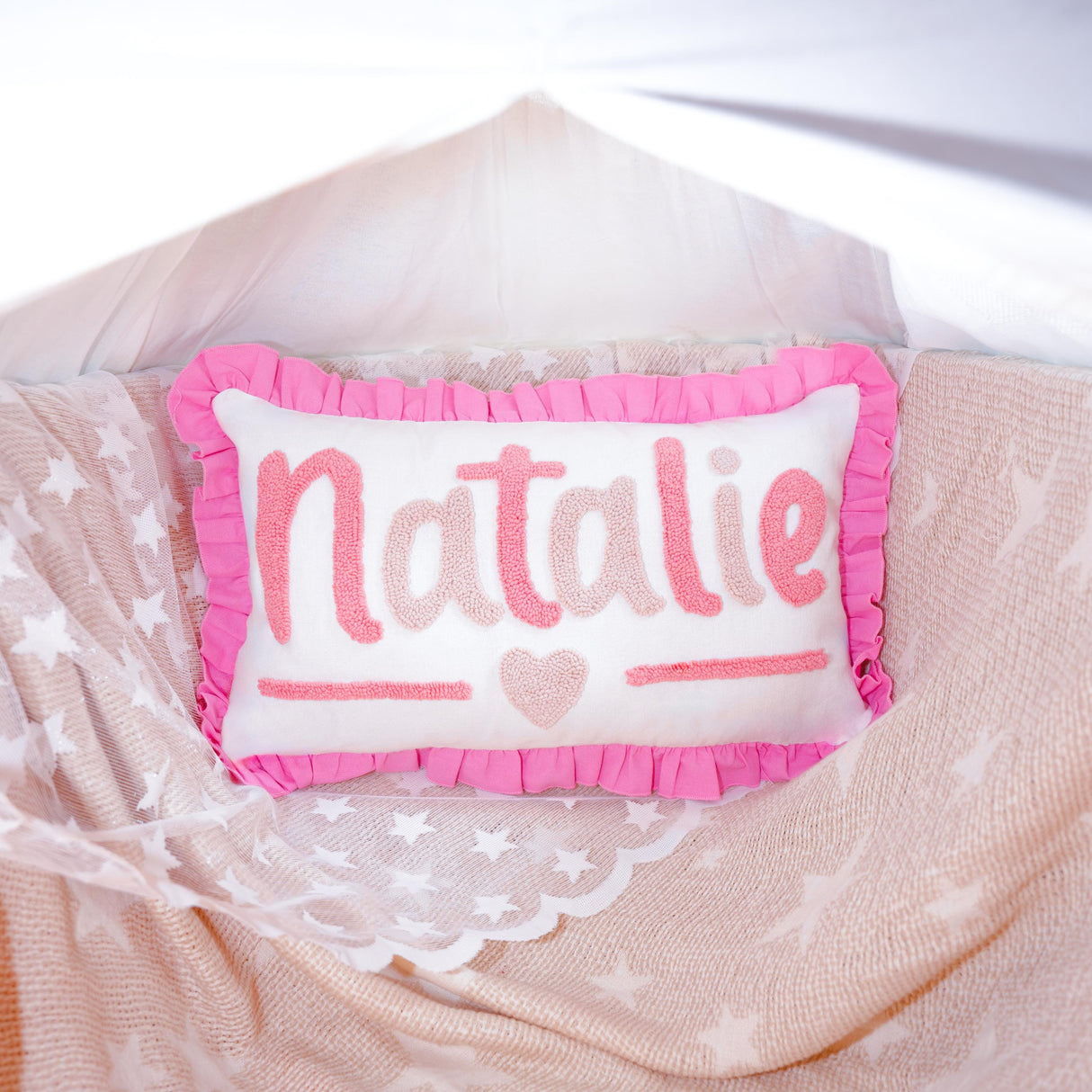 A pink and white pillow with the name &quot;NATALIE&quot; written on it, placed on a bed with a white sheet and a pink and white star-patterned blanket.