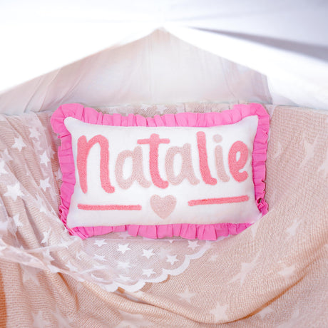 A pink and white pillow with the name &quot;NATALIE&quot; written on it, placed on a bed with a white sheet and a pink and white star-patterned blanket.