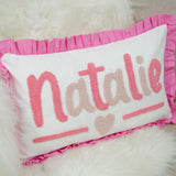 A white pillow with pink embroidered text and a pink ruffled border.