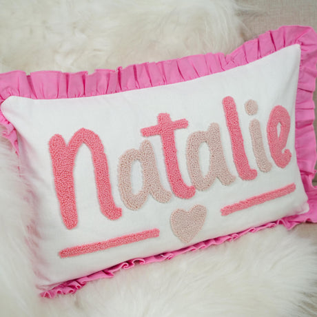 A white pillow with pink embroidered text and a pink ruffled border.