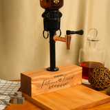 Wooden beer pump with engraved text on a wooden block, next to a glass decanter with liquid.