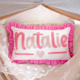 A pink and white decorative pillow with the name &quot;NATALIE&quot; written on it, placed on a bed with a white lace blanket.