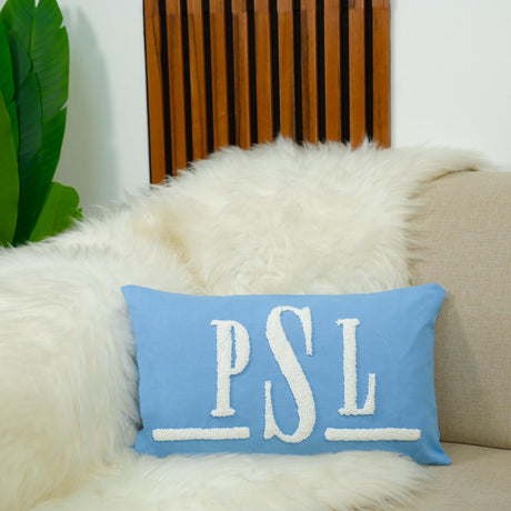 A blue pillow with the initials &quot;PSL&quot; on it is placed on a white couch, with a wooden wall and a green plant in the background.
