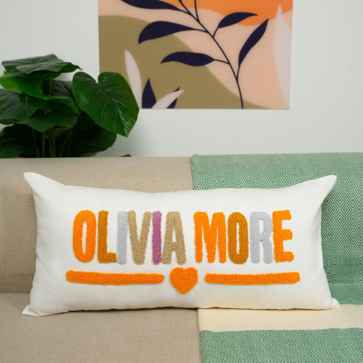 A white pillow with the text &quot;OLIVIA MORE&quot; in orange and pink colors, placed on a couch.