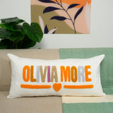 A white pillow with the text &quot;OLIVIA MORE&quot; in orange and pink colors, placed on a couch.