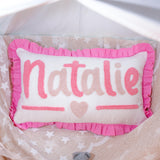 A pink and white pillow with the name &quot;NATALIE&quot; embroidered on it, placed on a bed with a white and beige patterned blanket.