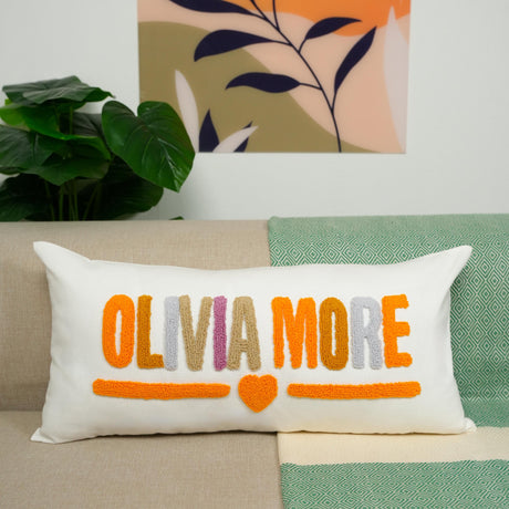 A white pillow with the text &quot;OLIVIA MORE&quot; in orange and pink colors, placed on a couch.