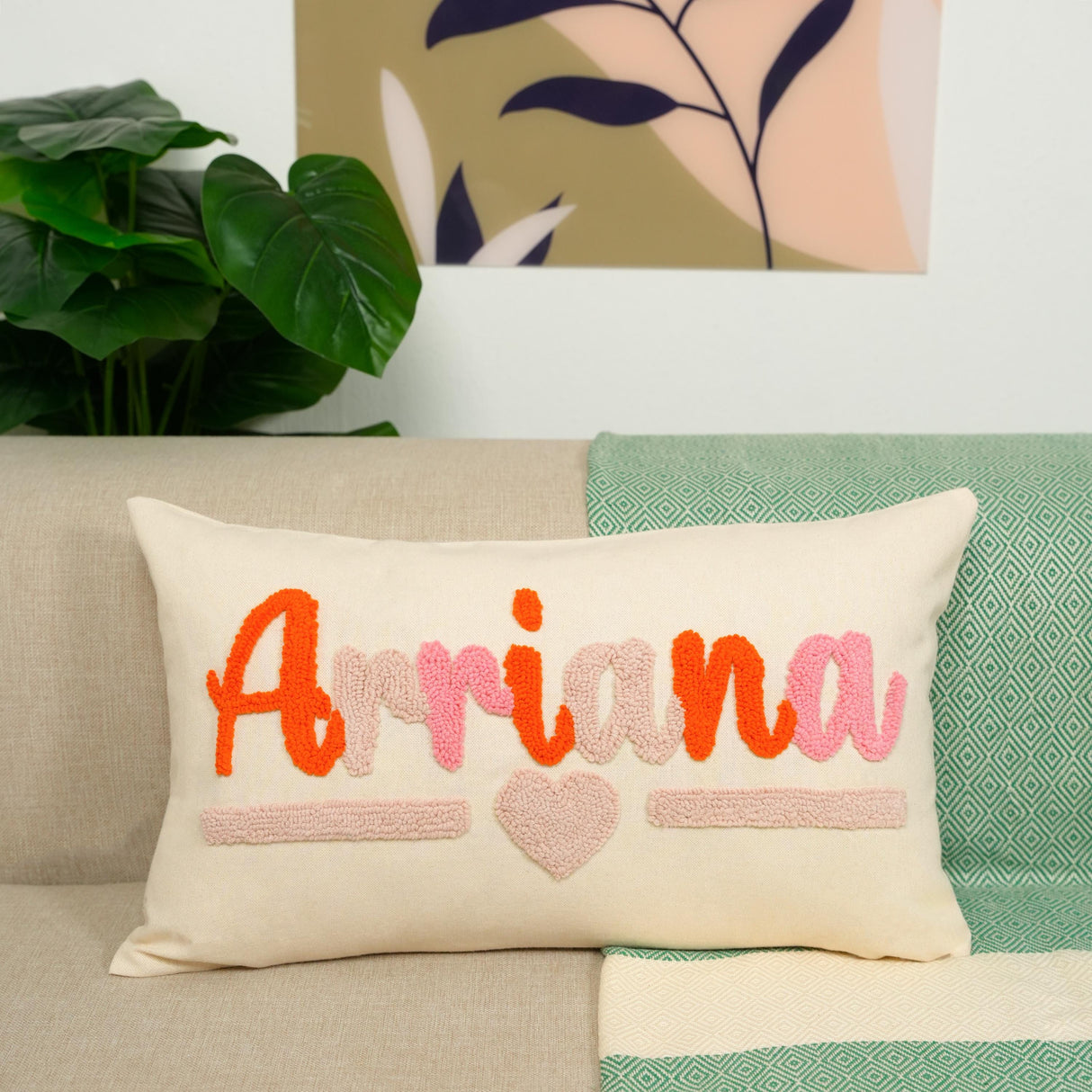 A beige pillow with the name &quot;Arriana&quot; and a heart design sits on a couch, with a green and white patterned blanket and a potted plant in the background.