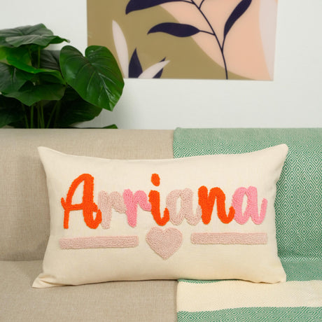 A beige pillow with the name &quot;Arriana&quot; and a heart design sits on a couch, with a green and white patterned blanket and a potted plant in the background.