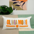 A white pillow with the text &quot;OLIVIA MORE&quot; in orange and pink colors, placed on a couch.